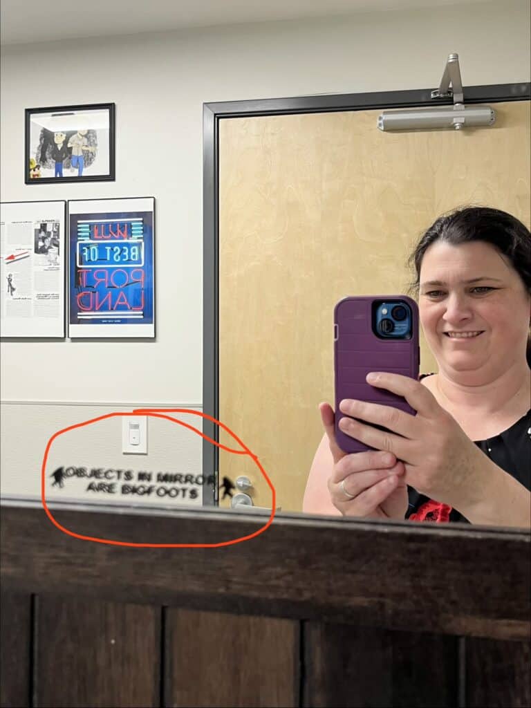 Jenn takes a mirror selfie. On the bottom left of the mirror is a sticker that reads "Objects in mirror are Bigfoots".