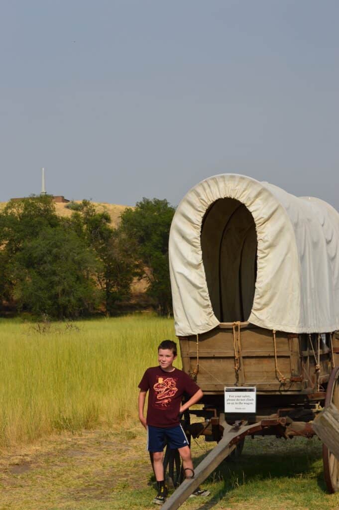 Our son stands next to an old covered wagon on the Oregon Trail ruts at Whitman Mission NHS.