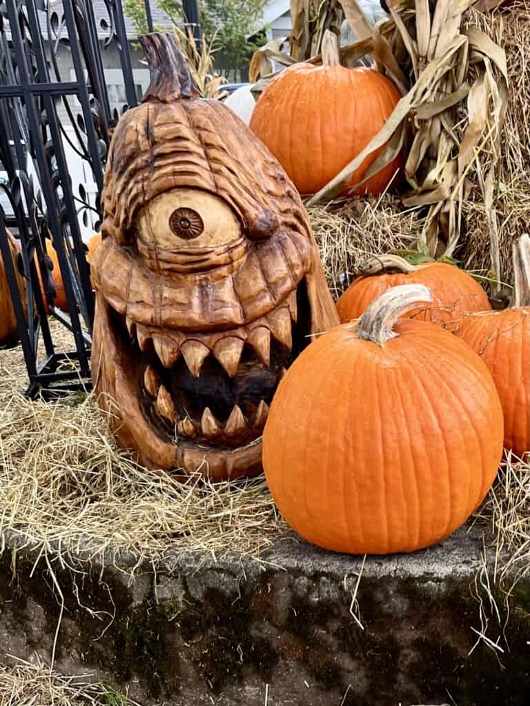 Orange pumpkins sit on a bed of hay with a giant one-eyed wooden jack-o-lantern next to them.