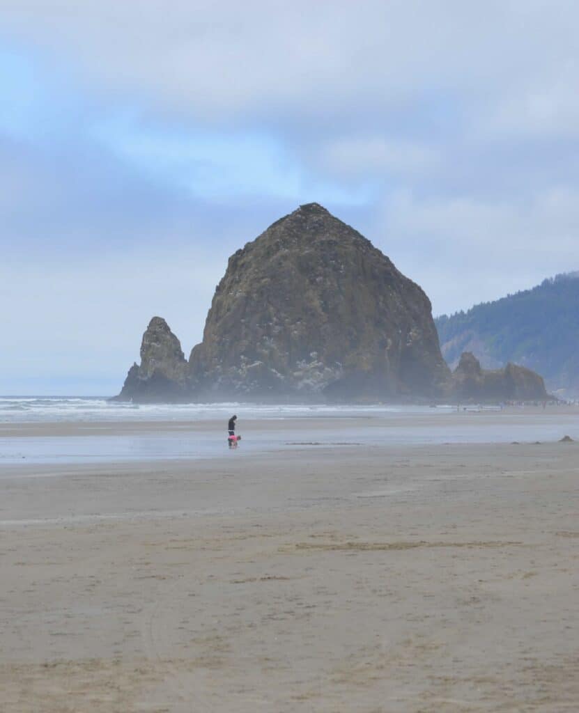 Haystack Rock towers over two pedestrians as they walk along Cannon Beach.