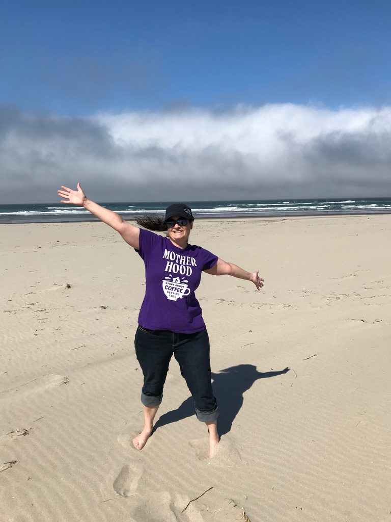 My wife stands with her arms stretched out and smiling in the sunshine at Sitka Sedge State Park.