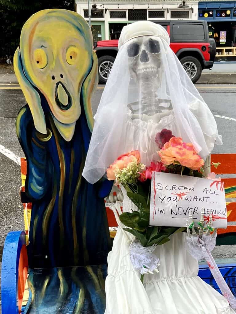 Colorful bench with a wood cutout of Mensch's "The Scream" person sits next to a skeleton bride holding a sign that says, "Scream all you want, I'm NEVER leaving!"