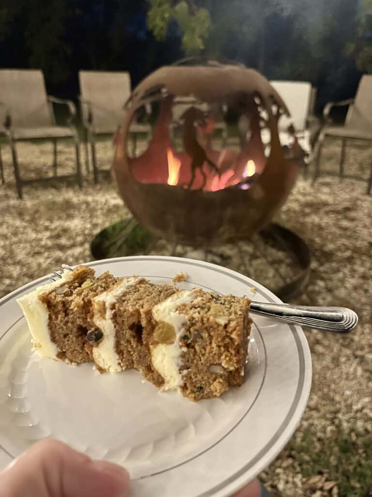 Carrot cake slice on a white plate with a fire pit in the background.